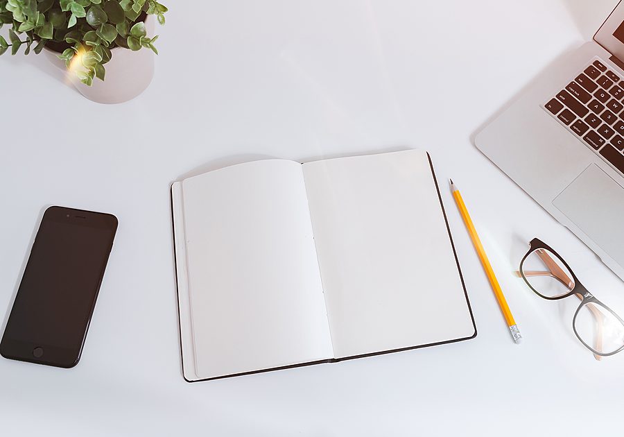 Notebook, laptop, phone and glasses laid out on a bright desk